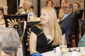 A woman with long blonde hair sits at a table and smiles at something off camera